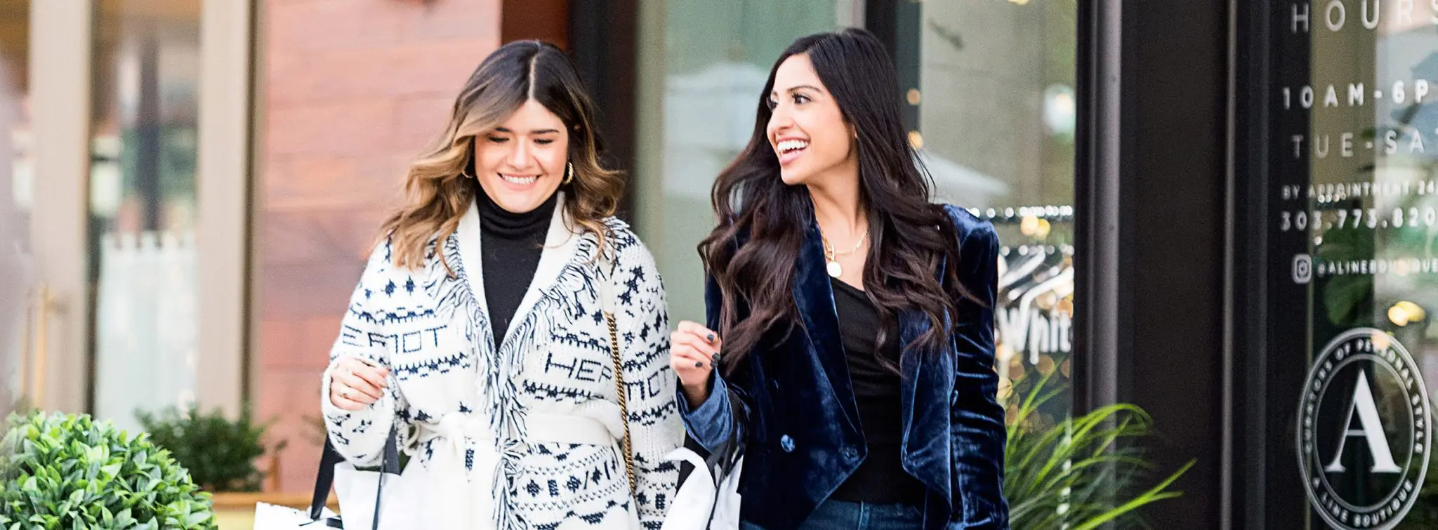Two smiling women shopping at a boutique with "HOURS" sign visible.