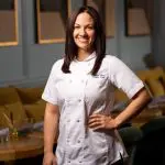 Pastry Chef Laura Kline in chef's coat, smiling in a restaurant. "Laura Kline, Pastry Chef" is embroidered on the coat.
