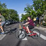 Father and son biking across a crosswalk on a sunny street, enjoying Father's Day 2024.