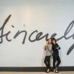 Two women pose by "Sincerely" wall art, NINE dot ARTS.