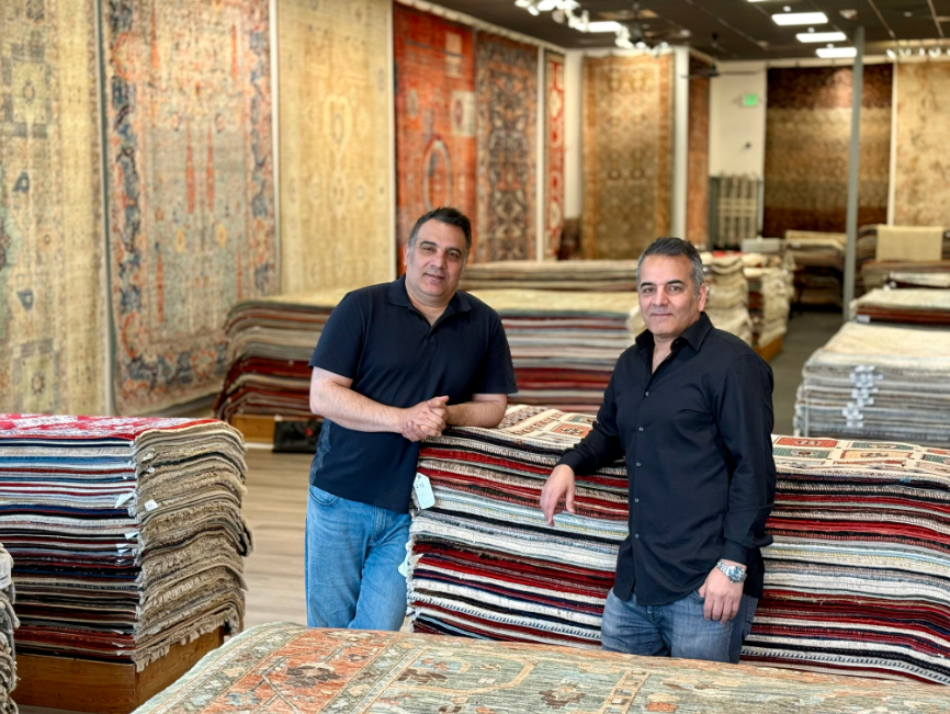 Kian Rugs Co. owners stand proudly in their rug store, surrounded by stacks of colorful carpets.