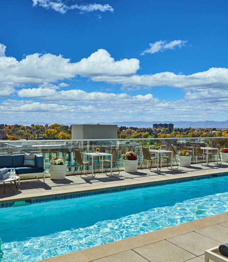 Rooftop pool with patio furniture and a city view under a blue sky.