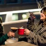 Two women toasting drinks at a rooftop bar; one taking a photo.
