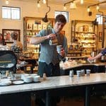Barista pouring coffee at Aviano Coffee, with other staff and customers in the background.