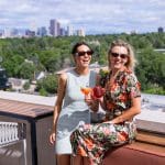 Two women toasting drinks on a Cherry Creek rooftop with Denver skyline views.