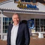 Man in suit stands in front of Déjà Blue Boutique. Store sign reads "déjà blue BOUTIQUE.