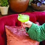 Green cocktail with cucumber garnish on a red couch.