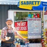Man at Henry's Hot Dogs cart, holding a hot dog. Menu visible, including Sabrett and Polish sausage.