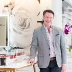 Man in a gray blazer standing in a makeup studio with lipsticks and orchids.