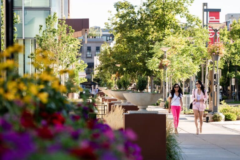 Two women walk on a tree-lined sidewalk with flowers.