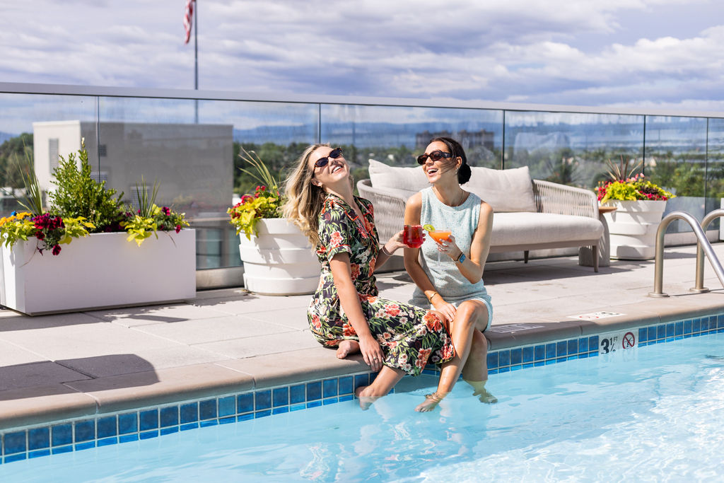 Two women enjoying cocktails poolside on a sunny rooftop.