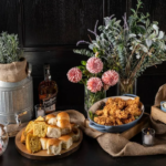 Fried chicken, dinner rolls, and whiskey on a rustic table setting.