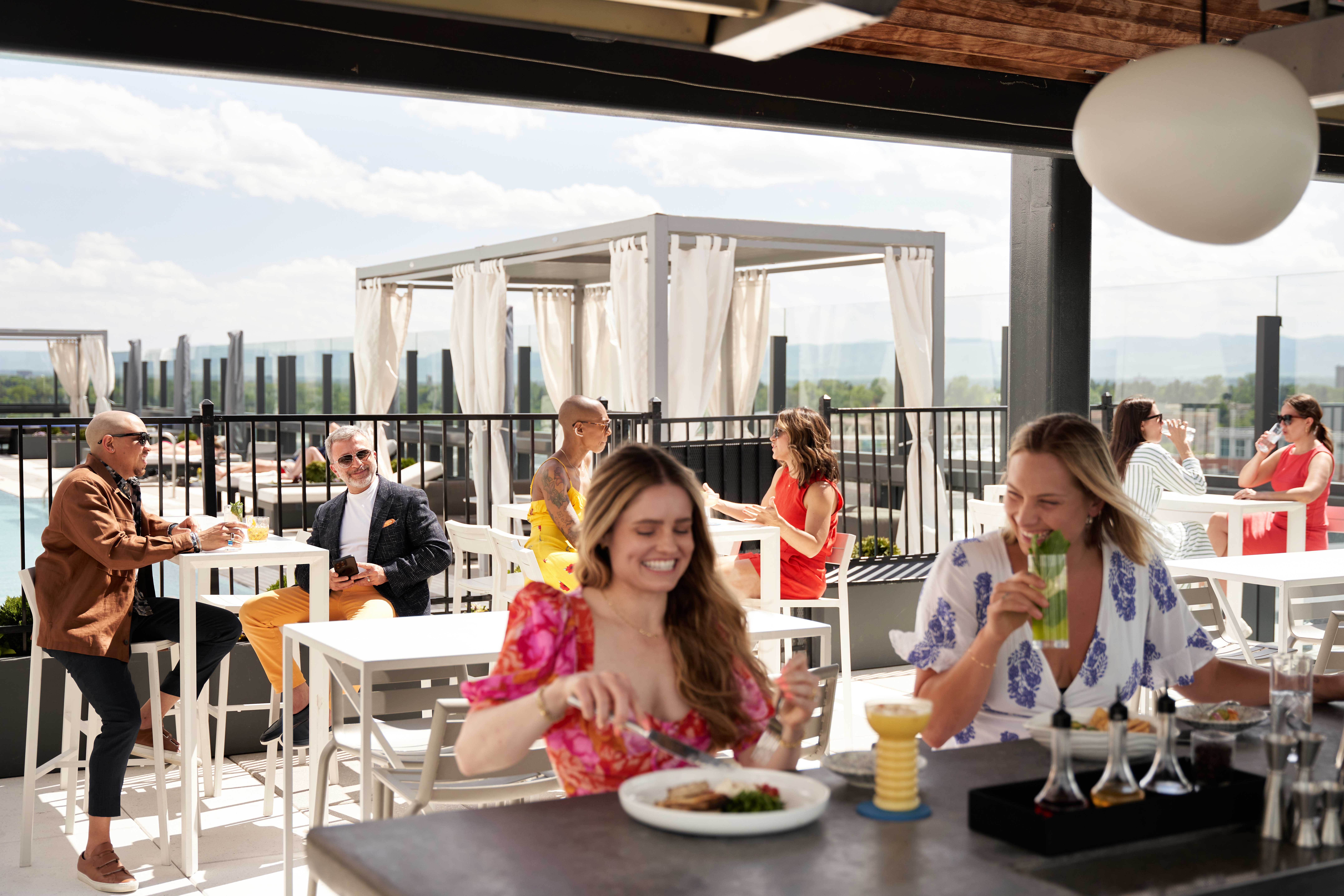 People dining on a rooftop patio with city views, enjoying food and drinks at tables and a bar.