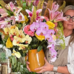 May floral class woman holds large floral arrangement with orchids and lilies in an orange vase