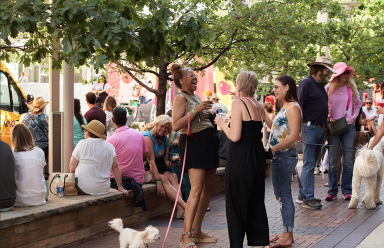 People enjoying an outdoor summer event under trees, with dogs and a yellow bus in the background.