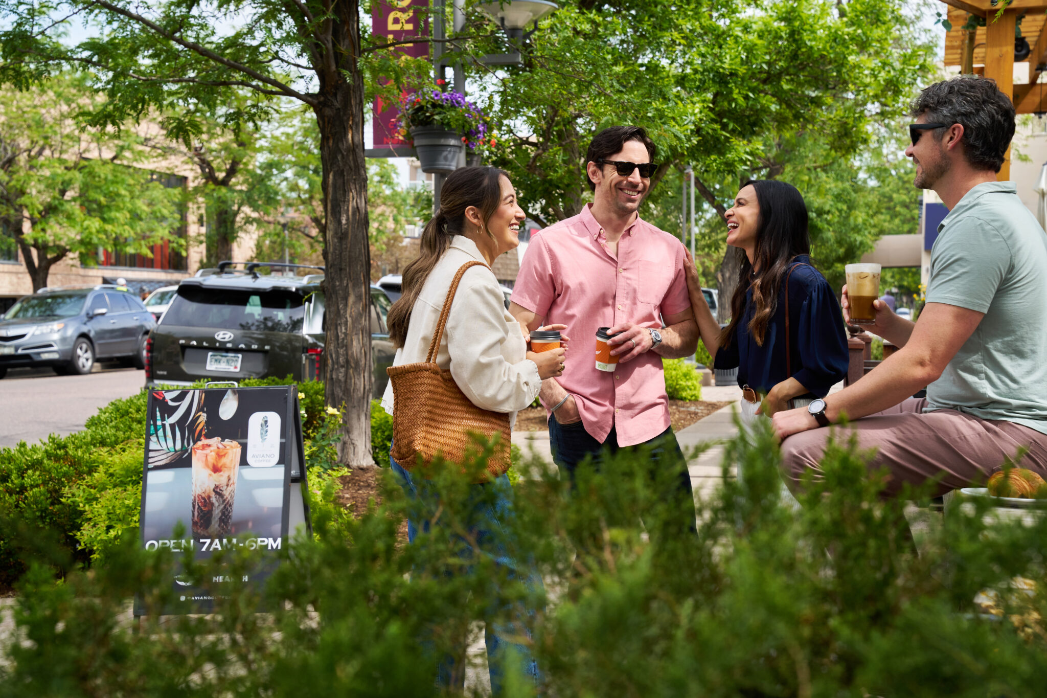 Four friends in casual clothes laugh together on a sunny sidewalk, holding coffee cups